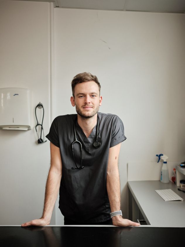 A man wearing dark grey scrubs standing behind a desk in a vet surgery, with a stethoscope around his neck