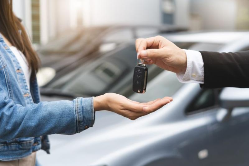 Someone handing a car key to a woman, a car in the background. 