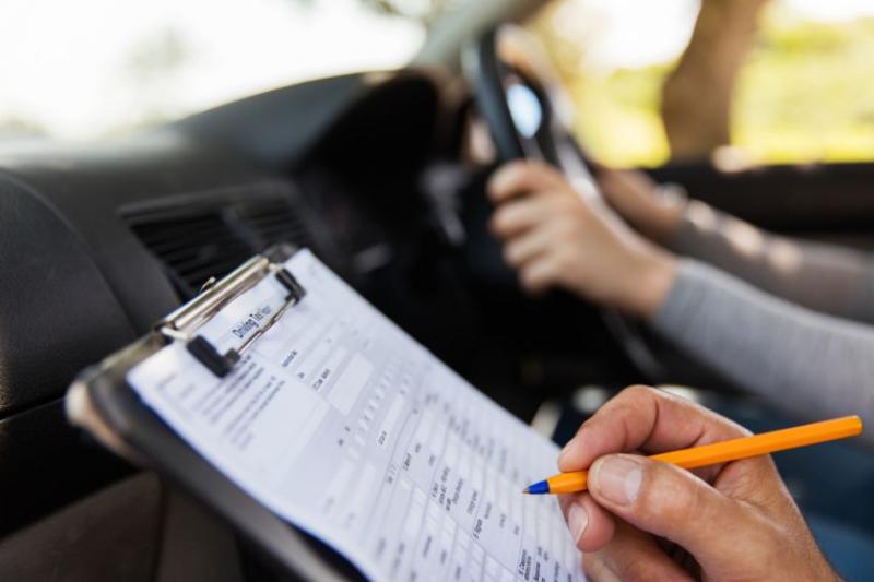 A view of a driving test clipboard with someone's hands on the steering wheel in the background