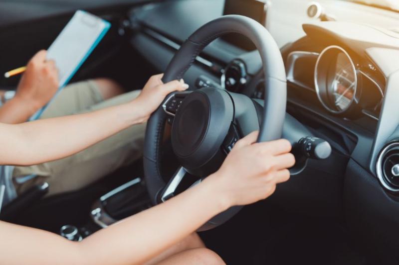 Two hands on steering wheel with someone making notes on clipboard in background