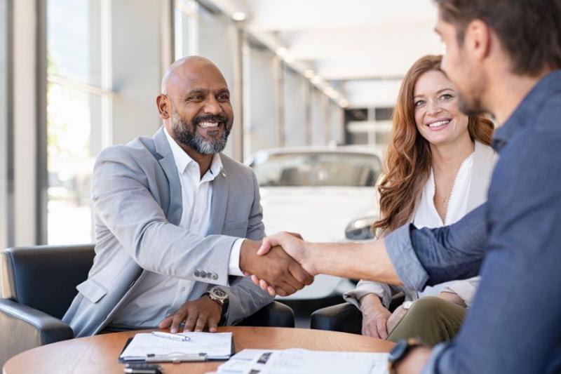 A man shaking hands with another man in a car dealership, a woman is sat beside them