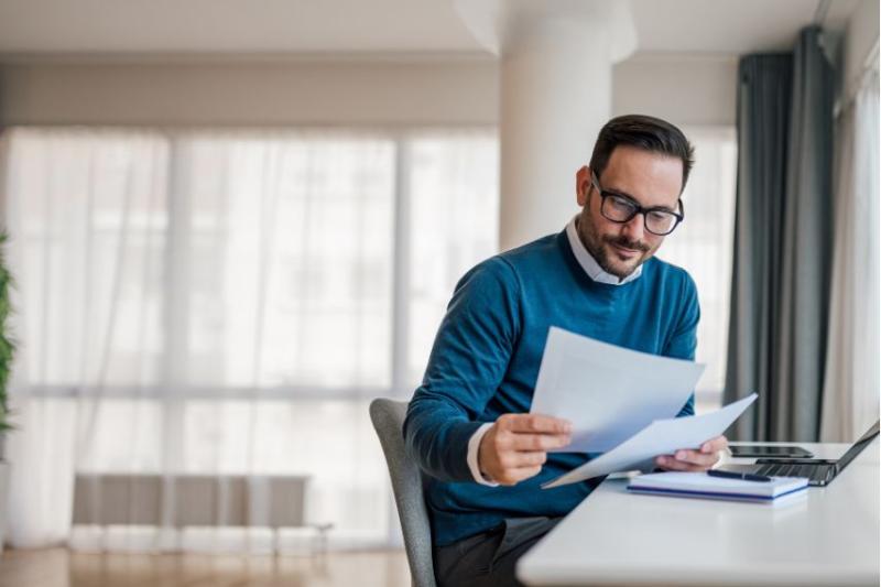 A man in a blue jumper and glasses reading papers in front of his laptop
