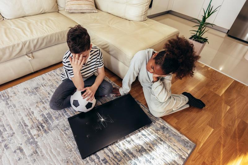 Sad children standing in front of a TV with broken screen holding a ball. Home insurance concept