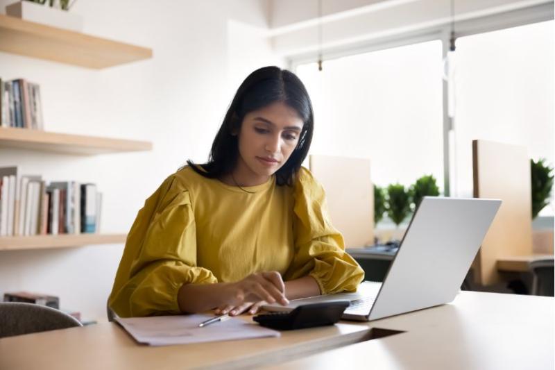 A woman in a yellow blouse using a calculator on a desk in front of her laptop
