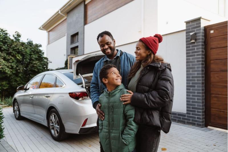 A mother, father and son stood in front of a silver car in front of a house