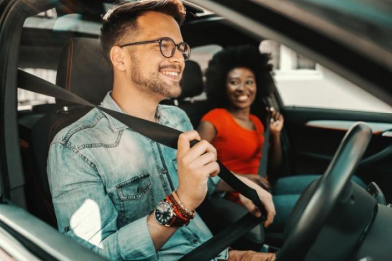 A man doing up his seatbelt in a car with a woman in a red top in the passenger seat