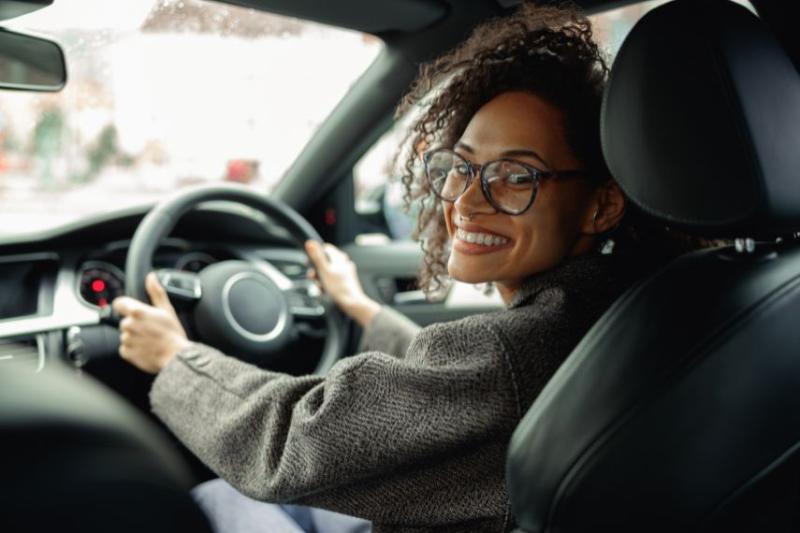 A woman smiling and wearing glasses, looking over her shoulder in the driver's seat of a car