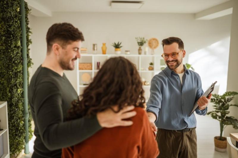 A smiling man in glasses and a shirt shaking hands with a couple in a flat