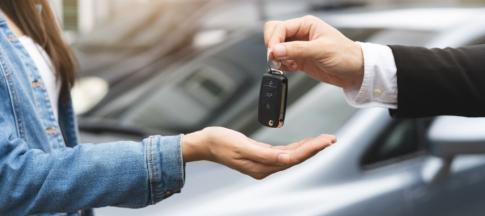 Someone handing a car key to a woman, a car in the background. 