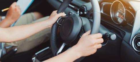 Two hands on steering wheel with someone making notes on clipboard in background