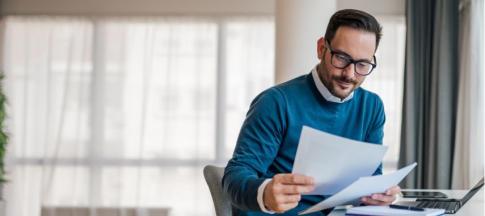 A man in a blue jumper and glasses reading papers in front of his laptop