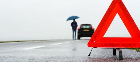 An orange triangle with a man broken down in his car in the background, with an umbrella