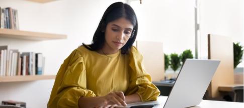 A woman in a yellow blouse using a calculator on a desk in front of her laptop