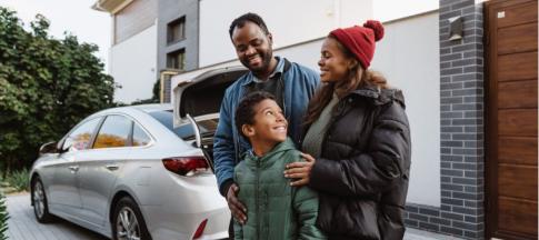 A mother, father and son stood in front of a silver car in front of a house