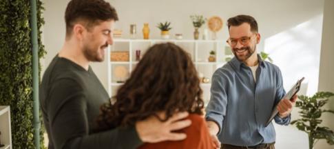 A smiling man in glasses and a shirt shaking hands with a couple in a flat