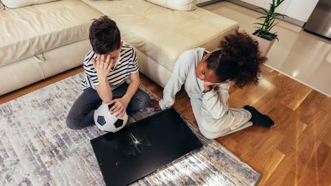 Sad children standing in front of a TV with broken screen holding a ball. Home insurance concept