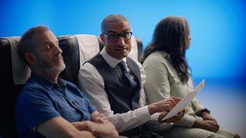 Airline attendant sitting between two Gen X passengers reading from airline safety sheet about pre existing medical conditions and travel insurance 